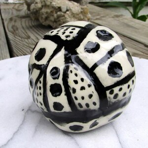 May include: A ceramic decorative sphere with a cream-colored base and a bold black geometric pattern. The design includes various shapes like dots, lines, and rectangles, creating a striking contrast. The sphere is displayed on a white surface.