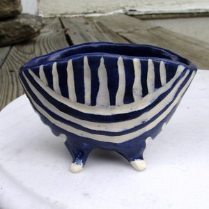 May include: A small, blue and white striped ceramic bowl with three legs. The bowl is hand-painted with white stripes on a dark blue background.