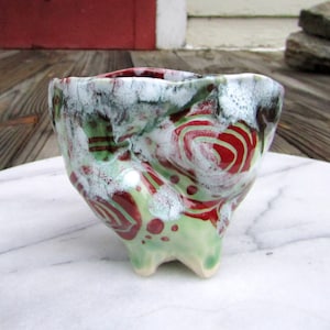 May include: A small, green ceramic bowl with red and white swirling designs. The bowl has three legs and a textured surface.