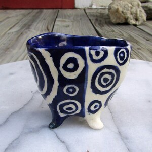 May include: A small, white ceramic bowl with three legs. The bowl is decorated with blue and white circles.
