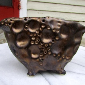 May include: A handcrafted, bronze-colored ceramic bowl with a textured surface and numerous small, circular perforations. The bowl has a unique, organic shape with three small feet and two protruding handles. The metallic glaze gives it a warm, earthy appearance.