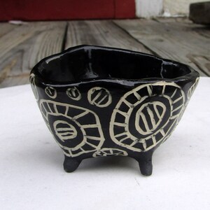 May include: A black ceramic bowl with three legs and a white geometric design. The bowl is decorated with circles, lines, and dots.
