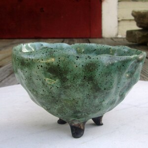 May include: A small, green ceramic bowl with a textured surface and three legs. The bowl has a speckled glaze and a slightly irregular shape.