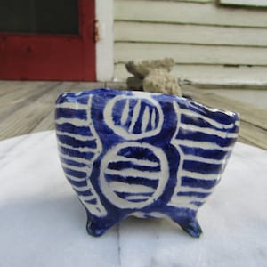 May include: A small, white ceramic bowl with three legs. The bowl is decorated with a blue and white geometric pattern.