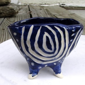 May include: A handmade ceramic bowl with a dark blue glaze and white spiral and dot pattern. The bowl has three small feet and a unique, irregular shape. The interior is a deep blue, and the rim has a slightly uneven edge.