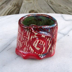 May include: A small, red ceramic cup with a textured, tree bark-like design. The cup has a green glaze on the inside and a white glaze on the outside.