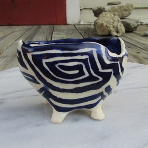 May include: A small, white ceramic bowl with three legs. The bowl is decorated with a blue and white geometric pattern.