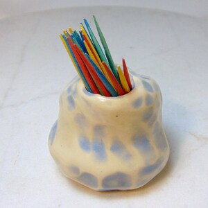 May include: A white ceramic toothpick holder with blue speckles. The holder is shaped like a small, rounded volcano and has a hole in the top for toothpicks. Multicolored toothpicks are sticking out of the holder.