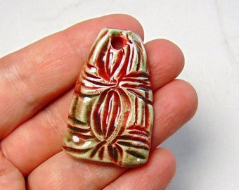 May include: A ceramic pendant with a red and green glaze. The pendant features a floral design and a hole at the top for hanging.