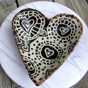 May include: A heart-shaped ceramic bowl with a black and white geometric pattern. The bowl is decorated with a repeating pattern of circles and hearts.