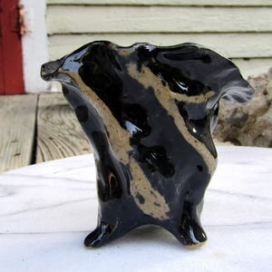 May include: A small, handcrafted ceramic vase with a unique, organic shape. The vase is primarily black with tan-colored, irregular stripes. It has a glossy finish and a ruffled top edge. The vase stands on three small feet.