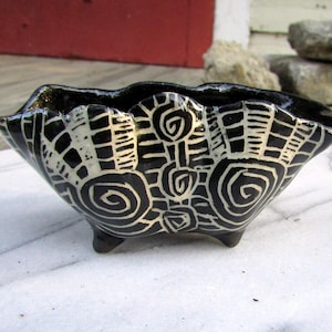 May include: A small, black ceramic bowl with a scalloped rim and three small feet. The bowl is decorated with a white, abstract pattern of spirals and geometric shapes. The bowl is sitting on a white surface.