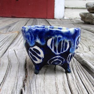 May include: A small, handmade ceramic bowl with a dark blue glaze and white abstract designs. The bowl has three small feet and a wavy rim. The exterior features a glossy finish. The bowl is placed on a weathered wooden surface.