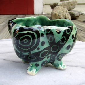 May include: A green ceramic bowl with black and white abstract designs. The bowl has three legs and a rounded shape.