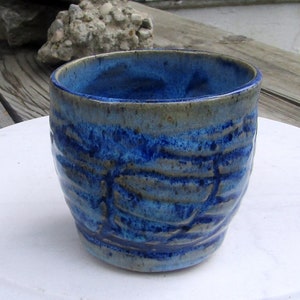 May include: A handmade ceramic cup with a textured, wave-like pattern. The cup features a deep blue glaze on the interior and exterior, with hints of lighter blue and brown. The cup is approximately 3.5 inches tall and 3 inches in diameter, perfect for coffee or tea.