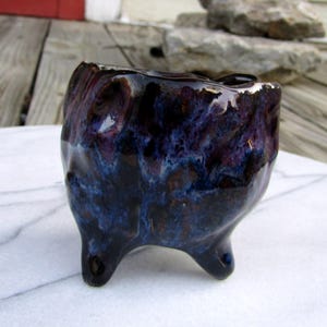 May include: A small, handmade ceramic pot with a dark, glossy glaze. The pot is primarily dark blue and purple, with a textured surface and three small, rounded feet. The rim has a lighter, almost white, edge.
