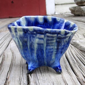 May include: A small, handmade ceramic bowl with a vibrant blue glaze. The bowl has a unique, textured surface with vertical streaks of lighter blue and white. It sits on three small feet and has a slightly irregular, wavy rim.