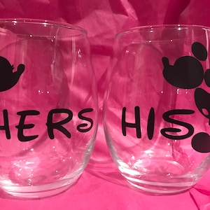 May include: Two clear glass wine glasses with black Mickey Mouse silhouettes and the words "Hers" and "His" printed on them.
