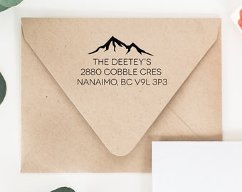 May include: A brown envelope with a black mountain range design and the text "THE DEETEY'S 2880 COBBLE CRES NANAIMO, BC V9L 3P3" printed on it. A wooden stamp with a red rubber mountain range design is also shown.