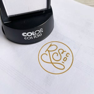 Op de afbeelding: Een zwarte zelf-inktende stempel met een gouden oneindigheidssymboolontwerp. De stempel is gelabeld "COLOP EOS R30". Een gouden ronde stempelafdruk van een cursief "R" met een hart erin bevindt zich op een wit oppervlak.
