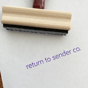 May include: A wooden stamp with a dark red handle and the words "return to sender co." stamped in purple ink on white paper. The stamp has a light wood block and a black ink pad.