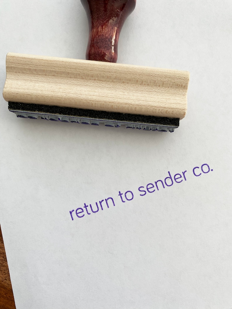 May include: A wooden stamp with a dark red handle and the words "return to sender co." stamped in purple ink on white paper. The stamp has a light wood block and a black ink pad.