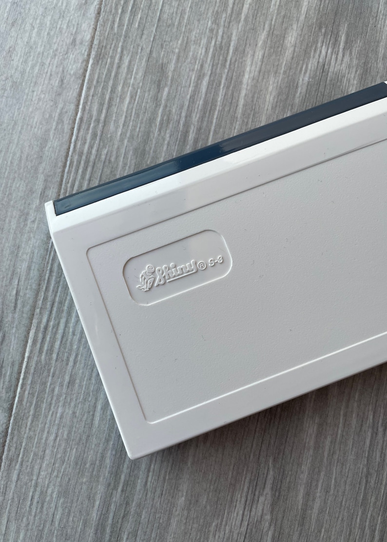 May include: A white rectangular object with a dark grey edge. The object has the embossed text "Smini S-3" in a stylized font. The object is on a light grey wooden surface.