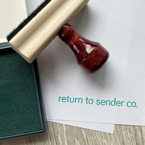 May include: A wooden stamp with a dark red handle and the text "return to sender co." printed in green. The stamp is next to a green ink pad.