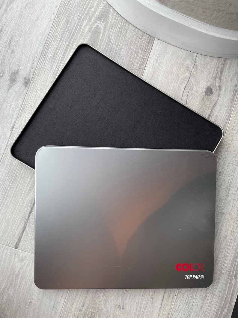 May include: A silver rectangular ink pad with rounded corners, displaying the text "COLOR TOP PAD" in red. The pad is open, revealing a black ink surface. The pad is on a light grey wooden surface.