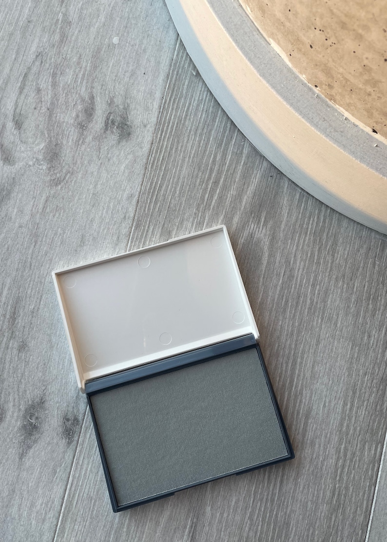 May include: An open, rectangular ink pad with a grey ink surface and a white lid. The ink pad is set against a light grey wooden surface, with a portion of a white and grey painted object in the background.