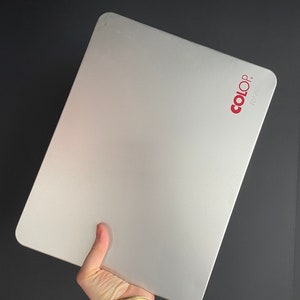 May include: A silver rectangular stamp pad with rounded corners. The brand name "COLOP" is printed in red in the upper right corner. The pad is held in a hand against a dark background.