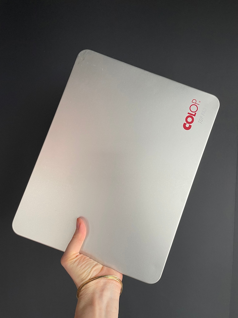 May include: A silver rectangular stamp pad with rounded corners. The brand name "COLOP" is printed in red in the upper right corner. The pad is held in a hand against a dark background.