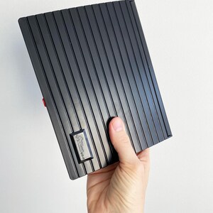May include: A black, rectangular item with vertical ridged lines, held in a hand. A small, rectangular emblem with text is visible on the item. The item has a red accent on the side. The background is plain white.