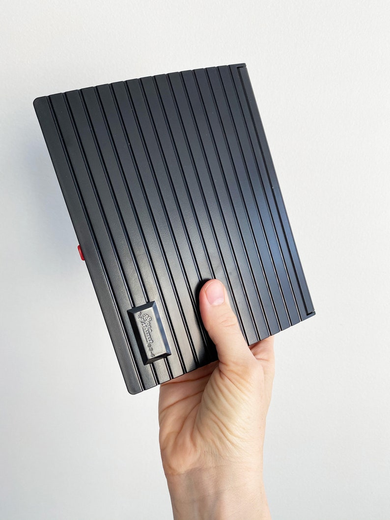 May include: A black, rectangular item with vertical ridged lines, held in a hand. A small, rectangular emblem with text is visible on the item. The item has a red accent on the side. The background is plain white.