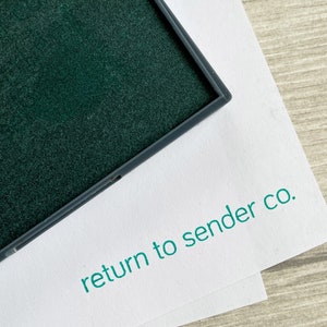 May include: A close-up of a dark green ink pad in a grey frame, on white paper. The words "return to sender co." are printed in teal on the paper. The background is a light wood grain surface.
