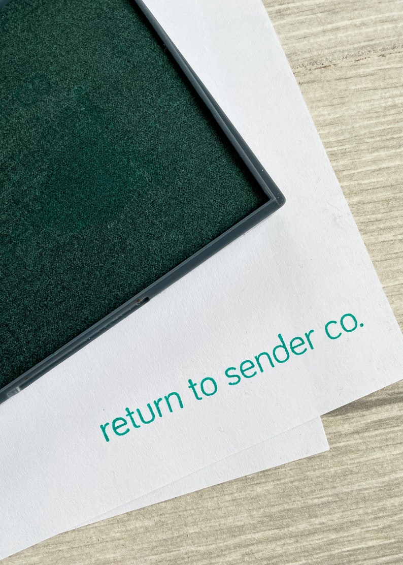 May include: A close-up of a dark green ink pad in a grey frame, on white paper. The words "return to sender co." are printed in teal on the paper. The background is a light wood grain surface.