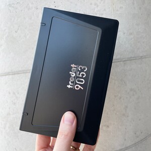 May include: A black rectangular stamp with the text "trodat 9053" in silver. The stamp is held by a hand. The stamp has a hinged lid and is likely used for office or crafting purposes.