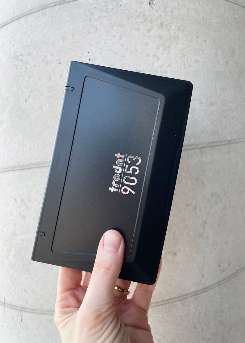May include: A black rectangular stamp with the text "trodat 9053" in silver. The stamp is held by a hand. The stamp has a hinged lid and is likely used for office or crafting purposes.