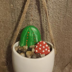 May include: A white ceramic hanging planter with a green and orange painted rock cactus and a red and white painted rock mushroom. The planter is filled with small brown and beige stones.