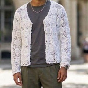 May include: A white open-front cardigan featuring an intricate crochet lace pattern with long sleeves and a scalloped edge, worn over a dark gray t-shirt and olive green trousers, accessorized with a silver chain necklace and a wristwatch.
