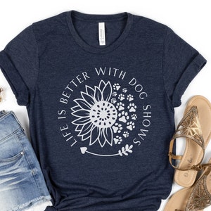 May include: A navy blue t-shirt with a white sunflower design. The sunflower has paw prints in place of petals. The text "Life is better with dog shows" is written around the sunflower.