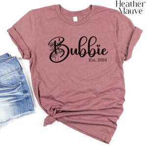 May include: Heather mauve t-shirt with the word "Bubbies" in a cursive font, with floral accents and a heart. The text "Est. 2024" is below the word "Bubbies". The shirt is knotted at the bottom.