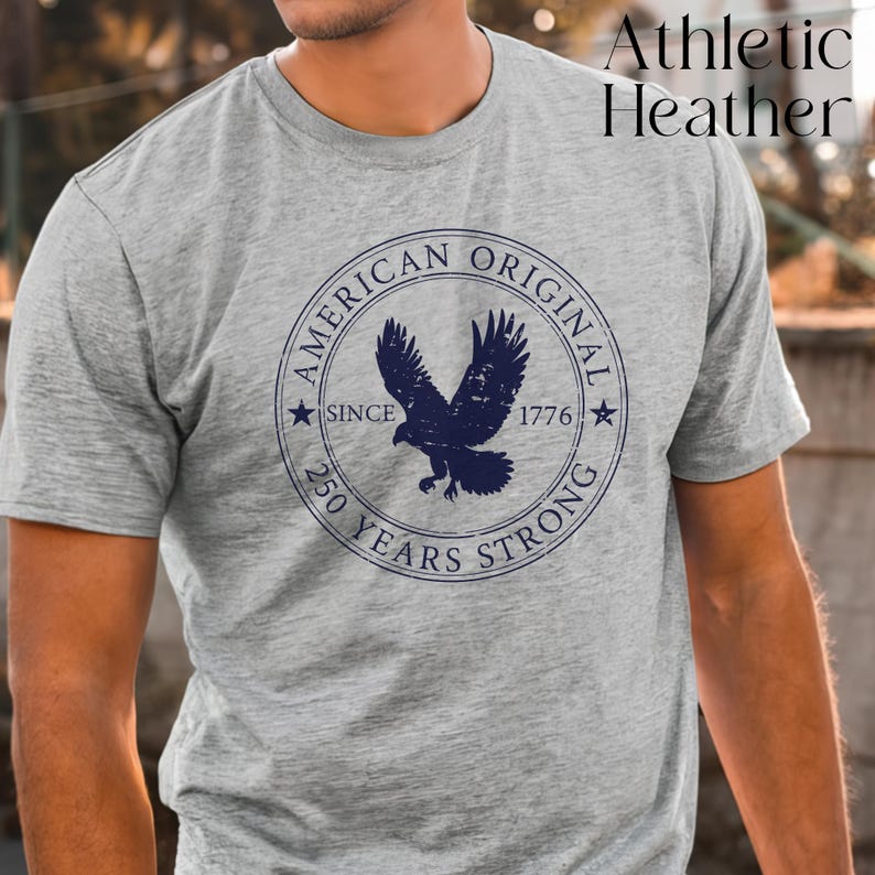 May include: Athletic Heather gray t-shirt with a navy blue circular graphic. The graphic features an eagle and the text "AMERICAN ORIGINAL SINCE 1776 250 YEARS STRONG". The shirt is short-sleeved.