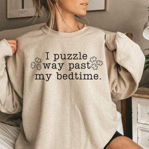 May include: A beige sweatshirt with the text "I puzzle way past my bedtime." and a graphic of puzzle pieces.