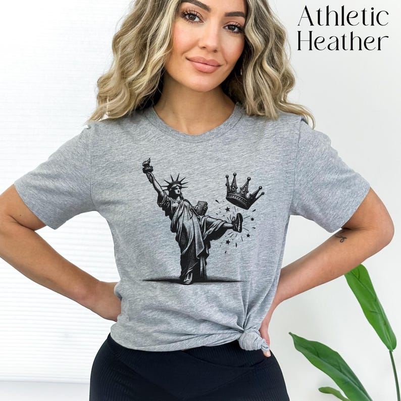 Może przedstawiać: Szara koszulka z czarno-białą grafiką przedstawiającą Statuę Wolności kopiącą koronę. Tekst "Athletic Heather" jest wydrukowany w prawym g&oacute;rnym rogu koszulki.