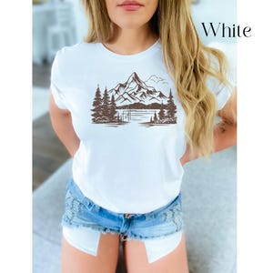 May include: White t-shirt with a brown mountain and lake graphic. The graphic features a mountain range with trees and a lake in the foreground.