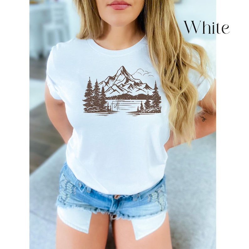 May include: White t-shirt with a brown mountain and lake graphic. The graphic features a mountain range with trees and a lake in the foreground.