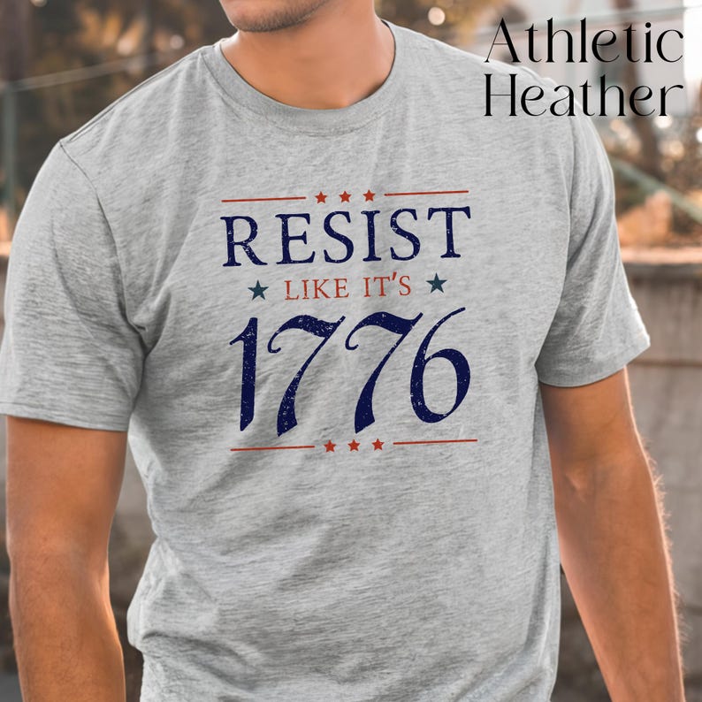 Pode incluir: Camiseta cinza mescla com o texto azul marinho e vermelho "RESIST LIKE IT'S 1776". O design inclui estrelas e linhas, criando um tema patri&oacute;tico. A camiseta &eacute; de manga curta.