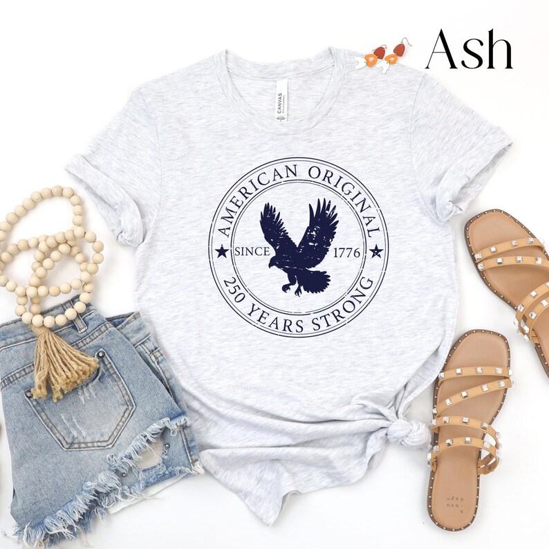 May include: Light gray t-shirt with a navy blue eagle graphic inside a circular design that reads "AMERICAN ORIGINAL SINCE 1776 250 YEARS STRONG." The shirt is paired with denim shorts and tan sandals.