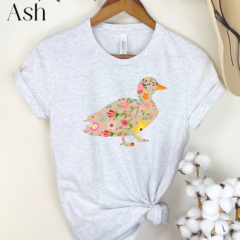 Peut inclure: Un t-shirt gris clair avec un motif de canard floral. Le canard est d&eacute;limit&eacute; par une ligne brune et rempli de fleurs color&eacute;es.